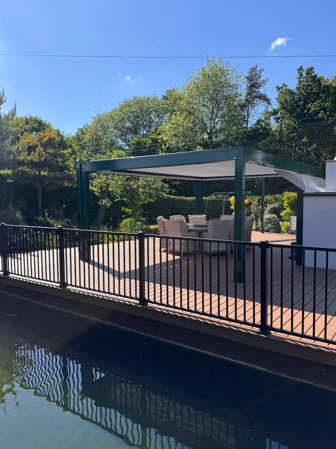 Black railing along a brown deck beside the water, a pergola behind