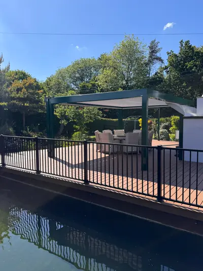 Black railing along a brown deck beside the water, a pergola behind