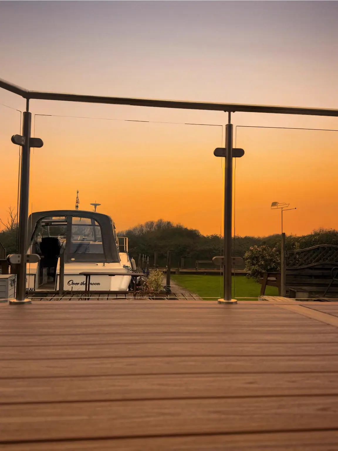 A glass balustrade with stainless steel posts on a brown deck, a boat moored beyond at sunset